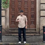 Young man wearing a beige shirt, black pants, and glasses standing outdoors in front of a wooden double door
