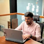 Man in a light pink shirt wearing glasses working on a laptop at a office desk